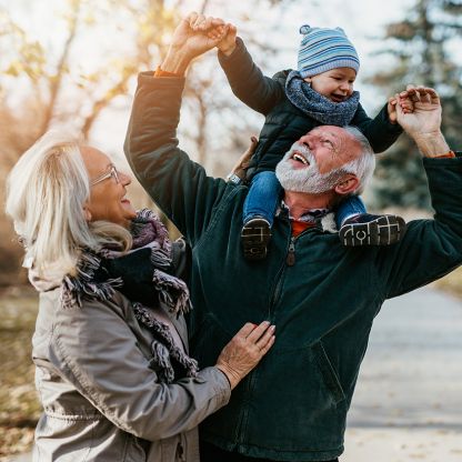 A man and a woman are smiling and looking up at their grandchild while the man carries the baby on his shoulders.