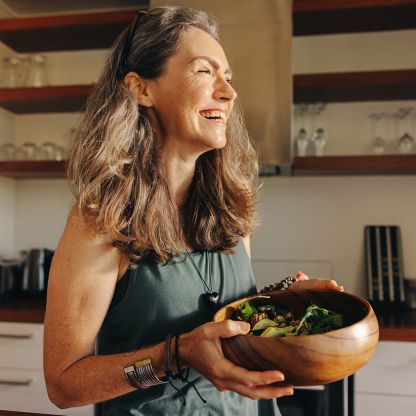 A woman is in the kitchen, holding a bowl with salad, and smiling.